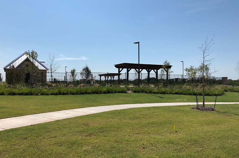 Grassy area with concrete path, brick building, wooden pergola, and metal fence