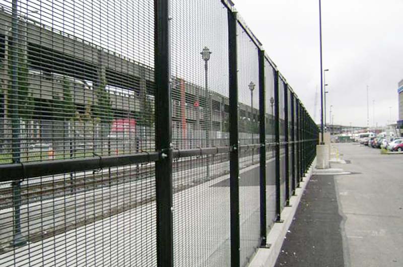 Black high-security fence enclosing train tracks and a parking structure. It has a tight mesh held on with ties to the top, middle and bottom rails. The posts are mounted on a concrete curb running the length of the fence.