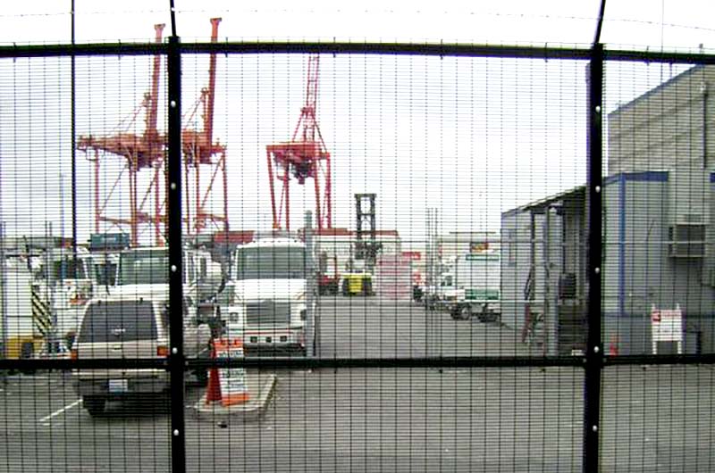 Black metal high-security fence enclosing an industrial shipping complex. It has a tight mesh held on with ties to the top and middle rails and is topped with barbed wire.