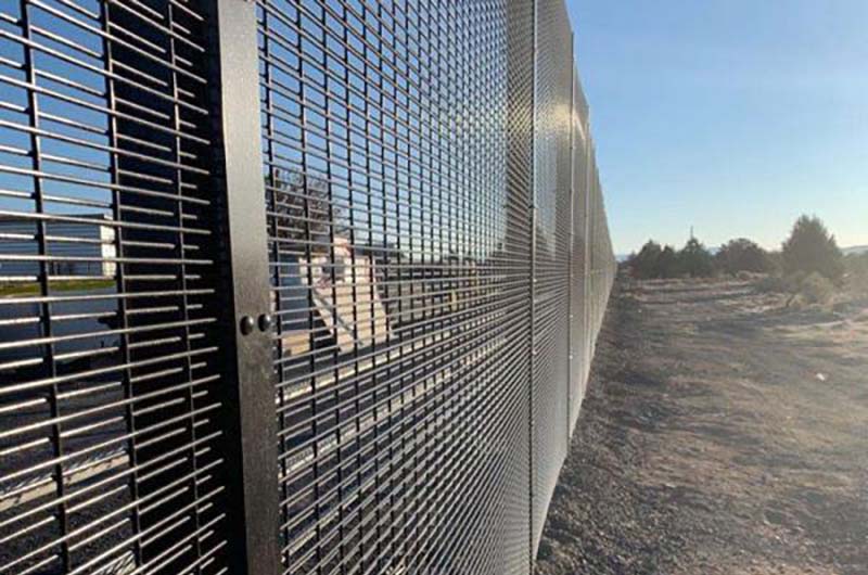 Tall, black metal high-security fence enclosing train tracks and industrial space. It has a tight mesh bolted onto the posts.