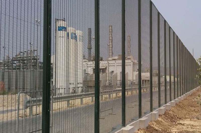 Tall, black metal high-security fence enclosing a road and industrial complex. It has a tight mesh bolted onto the posts and a concrete base running the length of the fence.