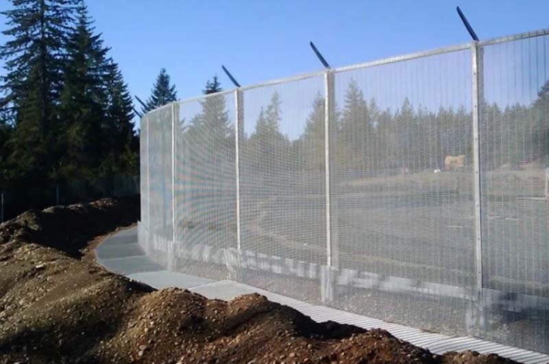 Tall, silver high-security fence enclosing a road and surrounded by trees. It has a tight mesh bolted onto the posts and a top rail with extensions that could hold barbed wire. Along the outer perimeter of the fence is a sidewalk and around the inner perimeter is a traffic barrier.